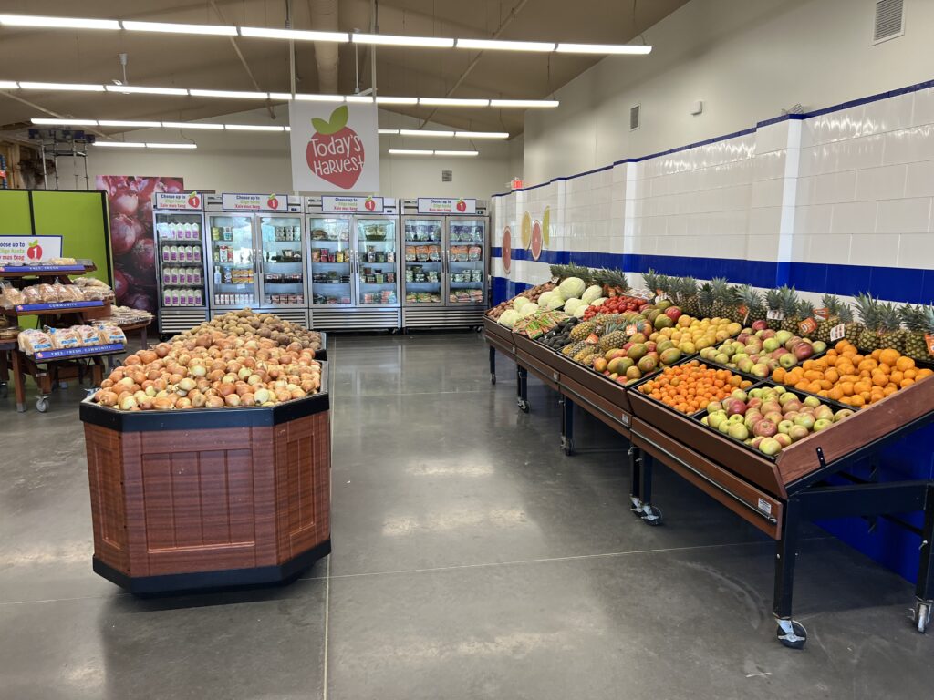 a shot of a Today's Harvest market without shoppers. To the left, a table with bakery items and a big produce bin. In the background, coolers filled with food, on the right, angled produce tables filled with a variety of food