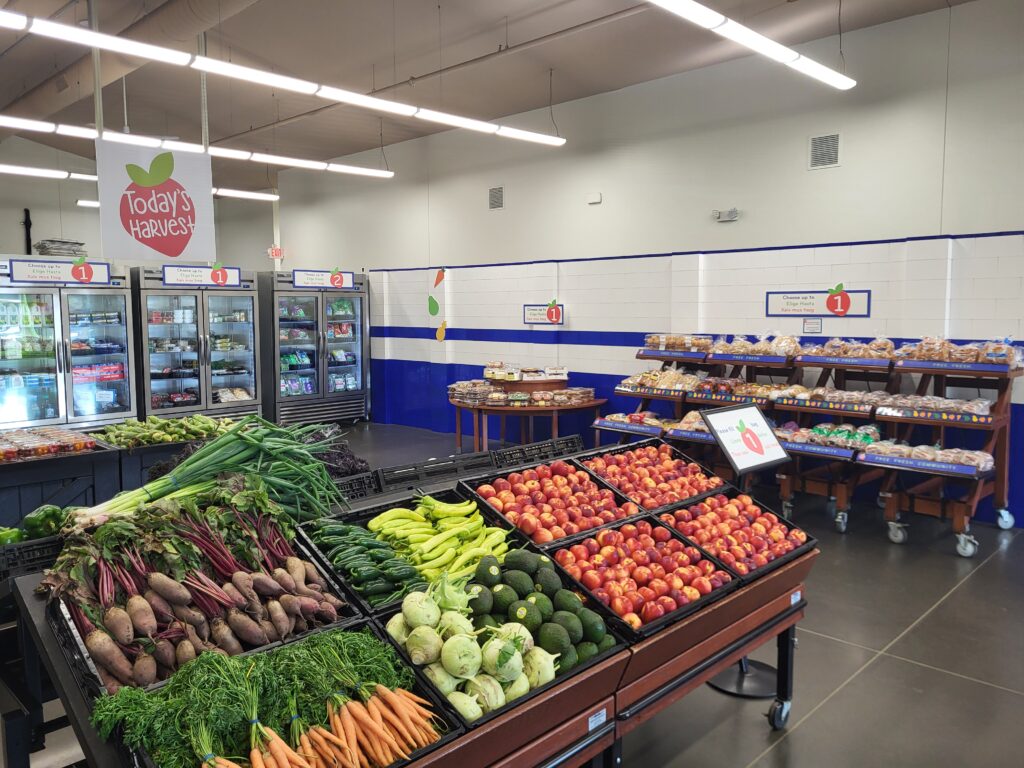 Today's Harvest market. In the foreground, there are angled tables with fresh produce including carrots, apples, and peaches. The background has food on wooden bakery racks and within fridges. You can see the logo of Today's Harvest within a strawberry hanging above the space.